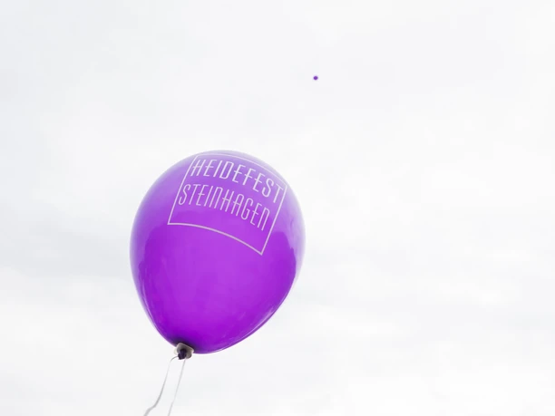 Ein einzelner lila Ballon mit der Aufschrift "Heidefest Steinhagen" schwebt vor wolkigem Himmel.