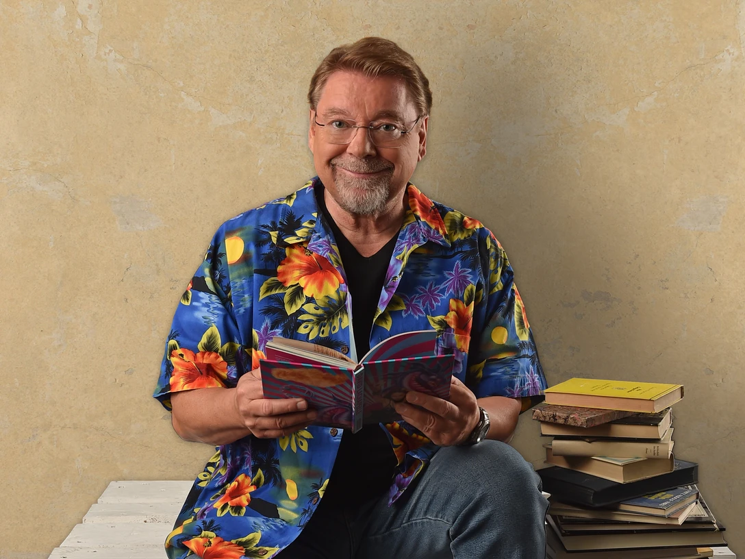 Ein lächelnder Mann in bunter, floraler Hemdbluse sitzt mit einem Buch und Bücherstapel vor hellem Hintergrund.