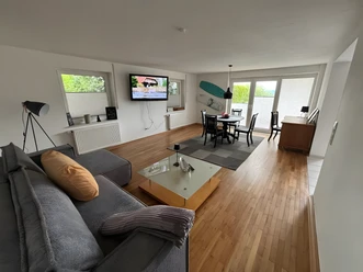Wohnzimmer Finca Bornrows Wohnzimmer mit Holzfußboden, grauem Sofa, Esstisch, Fernseher an der Wand und großen Fenstern.