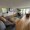 Wohnzimmer Finca Bornrows Wohnzimmer mit Holzfußboden, grauem Sofa, Esstisch, Fernseher an der Wand und großen Fenstern.