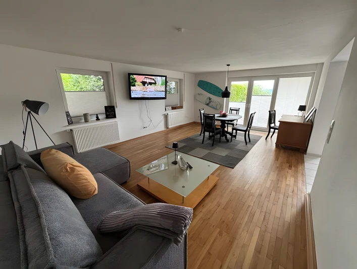 Wohnzimmer Finca Bornrows Wohnzimmer mit Holzfußboden, grauem Sofa, Esstisch, Fernseher an der Wand und großen Fenstern.