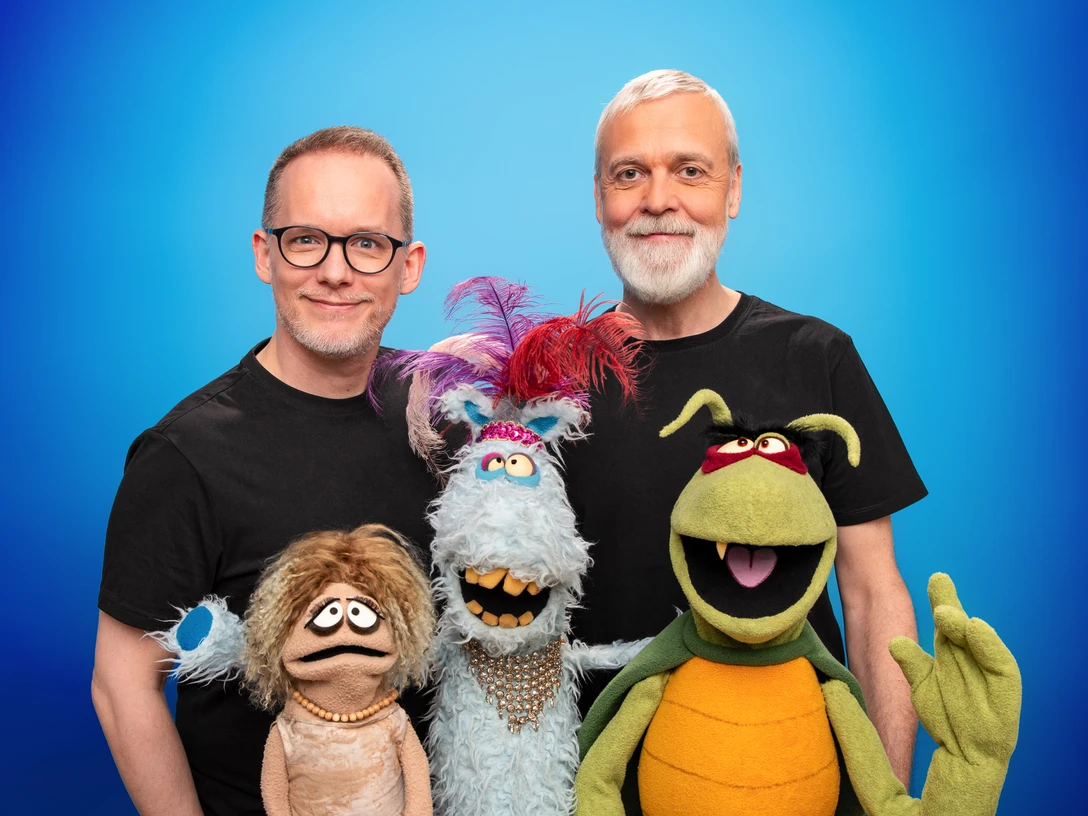 Puppen gucken 1_bigsmile2024(c)DannyFrede.jpg Zwei lächelnde Männer posieren mit bunten Puppen vor einem blauen Hintergrund.