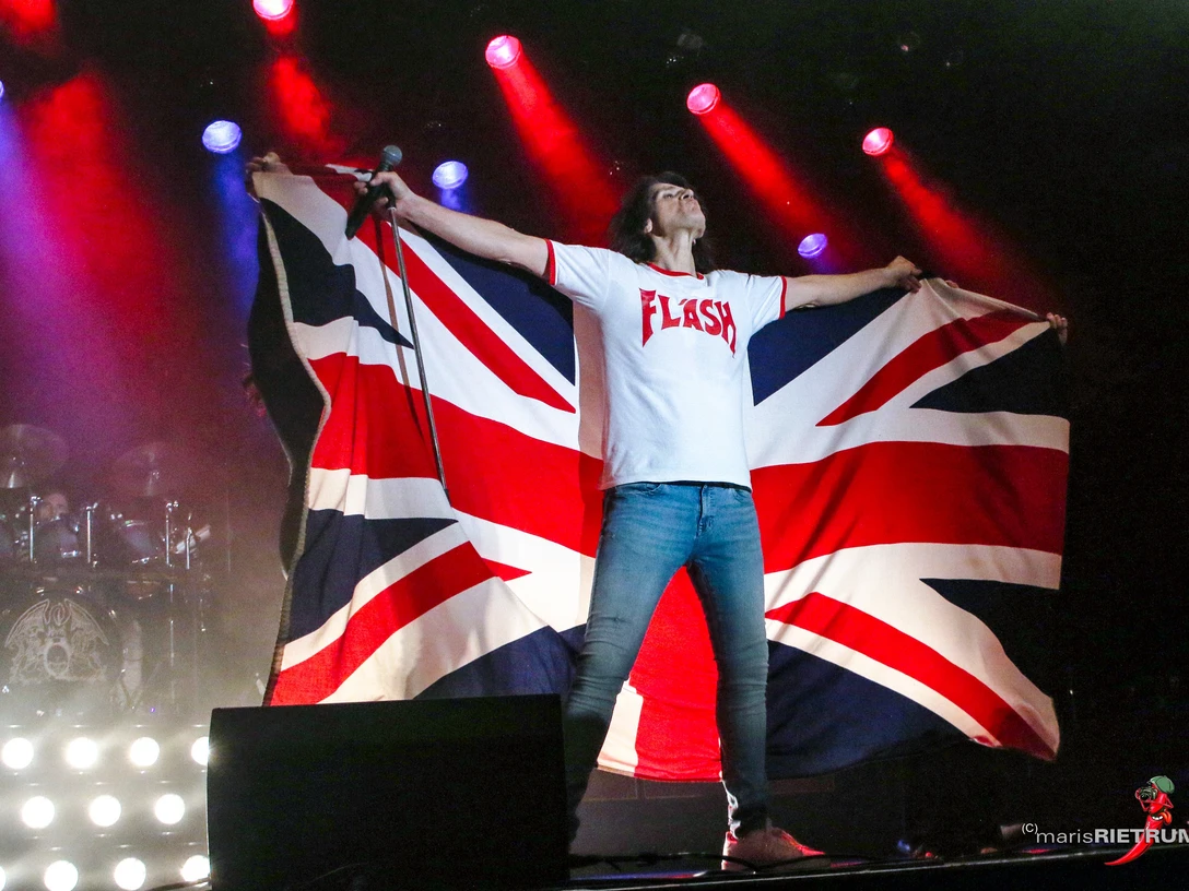 Ein Sänger in Flash-T-Shirt, hält eine Union Jack-Flagge auf der Bühne mit bunten Lichteffekten.