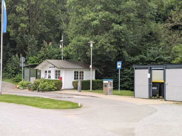 PXL_20240807_144221627.jpg Ein kleines weißes Gebäude neben einem Parkplatz, umgeben von grünen Bäumen und Sträuchern.