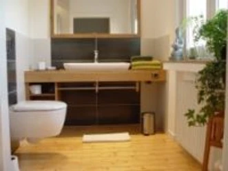 Modernes Badezimmer mit Holzfußboden, Wandwaschbecken, wandhängender Toilette und grünen Handtüchern.