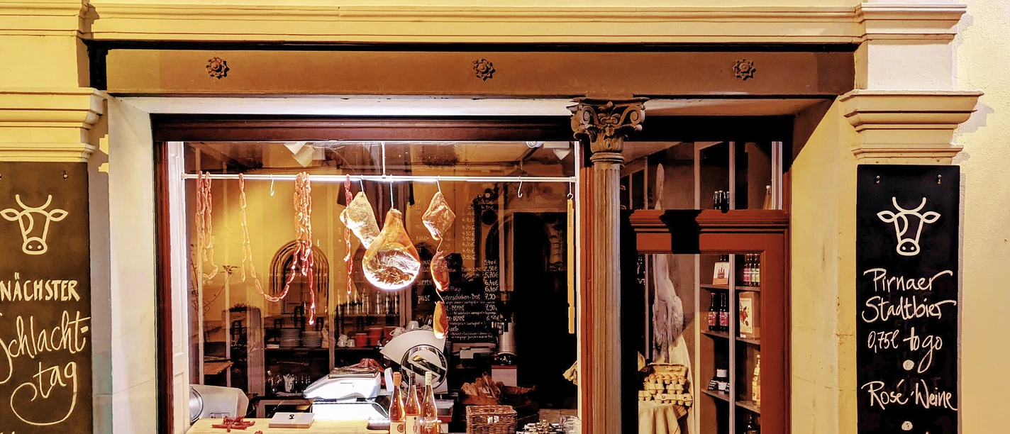 Hofladen15.jpg Fassade eines Hofladens bei Nacht, beleuchtet, mit Schaufenster voller Wurstwaren und Kreidetafeln mit handgeschriebenen Angeboten.