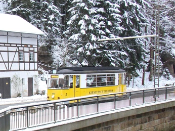 Kirnitzschtalbahn am Beuthenfall Eine gelbe Straßenbahn der Kirnitzschtalbahn fährt durch eine verschneite Waldlandschaft am Beuthenfall.A yellow Kirnitzschtalbahn streetcar travels through a snow-covered forest landscape at the Beuthen Falls.Žlutá tramvaj Kirnitzschtalbahn projíždí zasněženou lesní krajinou u Beuthenských vodopádů.Żółty tramwaj Kirnitzschtalbahn przejeżdża przez pokryty śniegiem leśny krajobraz przy wodospadzie Beuthen.Een gele Kirnitzschtalbahn tram rijdt door een besneeuwd boslandschap bij de Beuthen watervallen.Un tram giallo della Kirnitzschtalbahn attraversa un paesaggio forestale innevato presso le cascate di Beuthen.