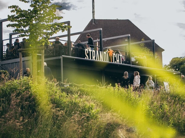 Ein moderner Außenbereich mit Besuchern vor dem Gebäude "Strandlicht" bei bewölktem Himmel.
