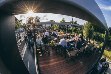 Menschen sitzen auf einer überdachten Holzterrasse und genießen bei Abendsonne Drinks und Gespräche.