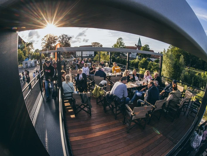 Menschen sitzen auf einer überdachten Holzterrasse und genießen bei Abendsonne Drinks und Gespräche.