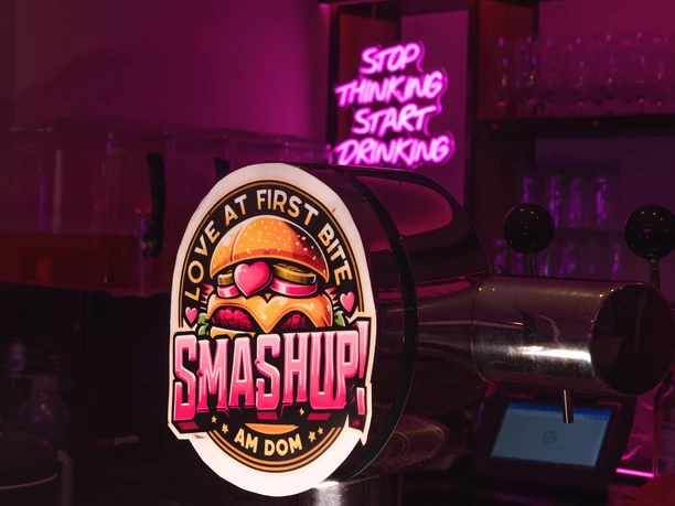 Theke mit einer Zapfanlage auf der "Smash u!" steht und Gläser im Hintergrund sowie die Leuchtschrift "Stop thinking start drinking".