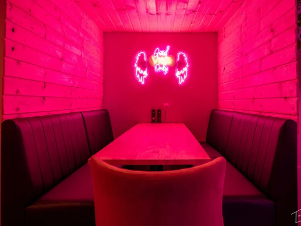 Pink beleuchteter kleiner Raum mit zwei Sitzbänken rechts und links und einem langen Tisch dazwischen. An der Wand hängt die Leuchtschrift "Good vibes only".