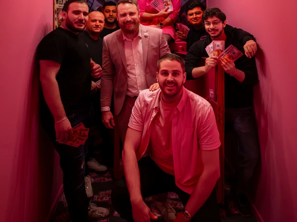 Smashup! 8.JPG Eine Gruppe von Personen steht in einem schmalen Raum mit Deckenlampe und Absperrseil und lächelt in die Kamera.