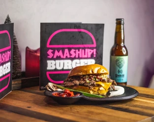 Tisch mit einem Burger auf einem Teller, einer Flasche Bier und einer aufgestellten Karte, auf der "Smash up Burger" steht.