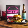 Smashup! 9.jpg Tisch mit einem Burger auf einem Teller, einer Flasche Bier und einer aufgestellten Karte, auf der "Smash up Burger" steht.
