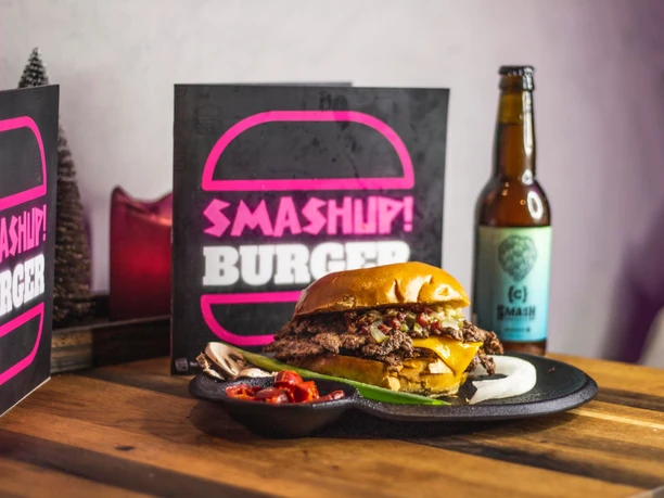 Smashup! 9.jpg Tisch mit einem Burger auf einem Teller, einer Flasche Bier und einer aufgestellten Karte, auf der "Smash up Burger" steht.