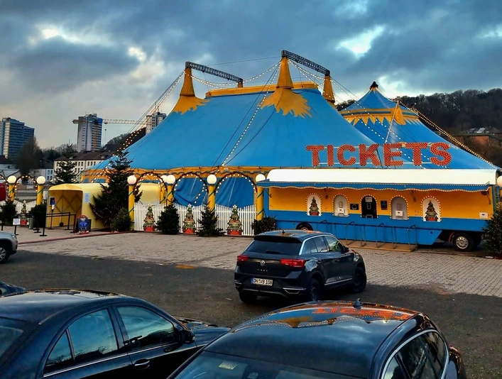 Weihnachtszirkus.jpg Ein großes blau-gelbes Zirkuszelt mit "TICKETS"-Schild, umgeben von Lichtern und Bäumen.