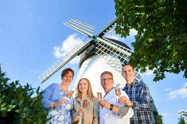 Vier Personen halten Gläser vor einer weißen Windmühle mit blauen Flügeln und lächeln.