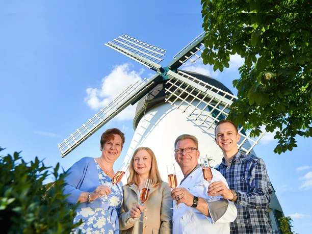 Vier Personen halten Gläser vor einer weißen Windmühle mit blauen Flügeln und lächeln.