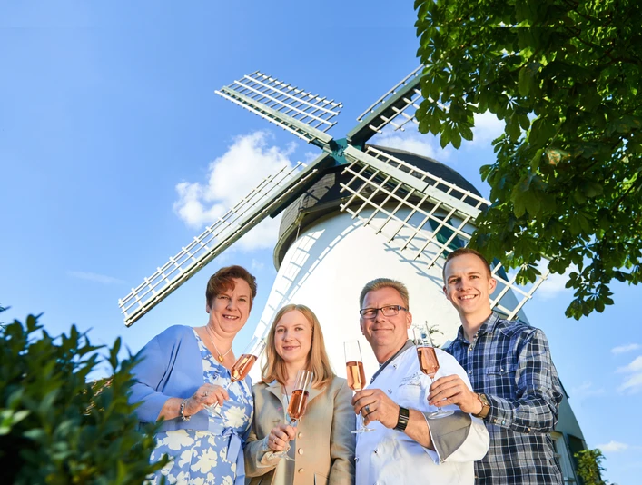Vier Personen halten Gläser vor einer weißen Windmühle mit blauen Flügeln und lächeln.