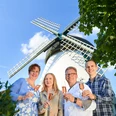 Bad Meinberg-Windmühle Fissenknick-Teutoburger Wald-LTM GmbH-Hendrik Lemke (2).jpg Vier Personen halten Gläser vor einer weißen Windmühle mit blauen Flügeln und lächeln.