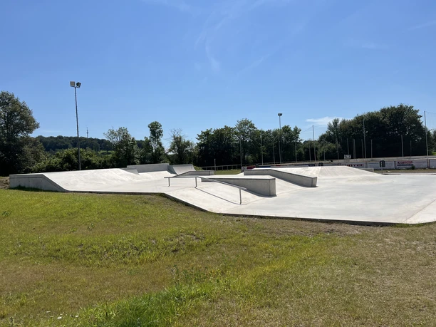 Skaterpark Much Moderner Skatepark mit Betonrampen und Geländern, umgeben von grünen Bäumen und Wiesen bei Sonnenschein.