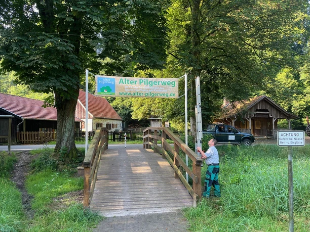 Ein Holzsteg führt zu einem Schild des Alten Pilgerwegs im Haxtergrund, umgeben von Bäumen.
