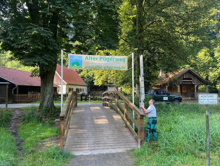 Ein Holzsteg führt zu einem Schild des Alten Pilgerwegs im Haxtergrund, umgeben von Bäumen.