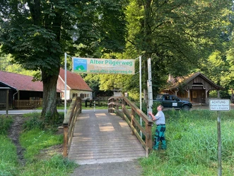 Banner Alter Pilgerweg im Haxtergrund Ein Holzsteg führt zu einem Schild des Alten Pilgerwegs im Haxtergrund, umgeben von Bäumen.