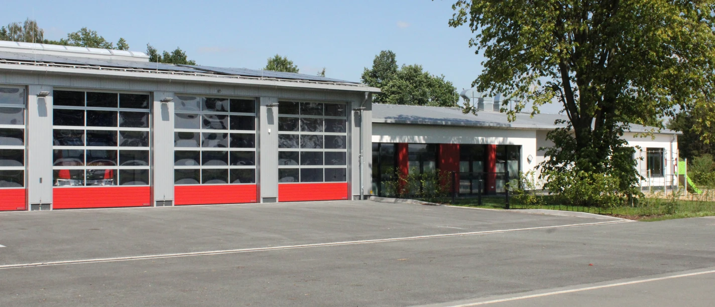Feuerwehrgerätehaus Lashorst 2024 Feuerwehrgerätehaus mit modernen roten Toren, befestigtem Parkplatz und angrenzendem Grünbereich.