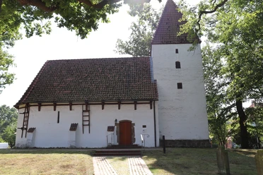 Weiße Kapelle mit markantem Satteldach und seitlichem Turm, umgeben von Bäumen und Natur.