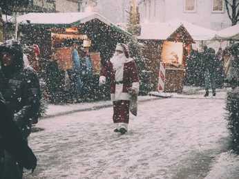Weihnachtsmann.jpg Ein Weihnachtsmann schlendert durch einen verschneiten Weihnachtsmarkt mit dekorierten Ständen.