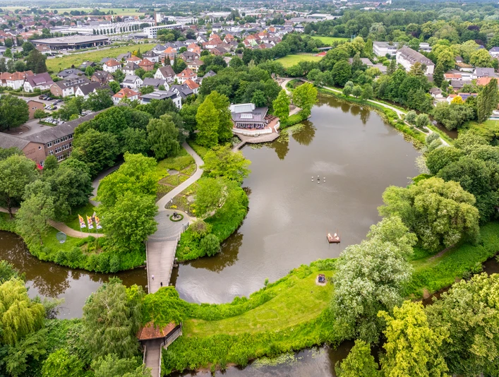 Luftaufnahme eines Parks mit See und Boot, umgeben von üppigem Grün und Stadtlandschaft im Hintergrund.