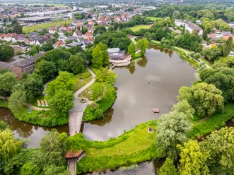 Rheda-Wiedenbrück-Emssee-Teutoburger-Wald-D-Ketz-094.jpg Luftaufnahme eines Parks mit See und Boot, umgeben von üppigem Grün und Stadtlandschaft im Hintergrund.