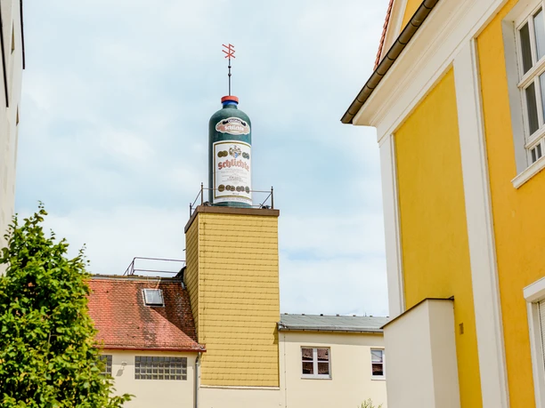 Schiltl-Getränketurm steigt zwischen gelben Gebäuden auf, blauer Himmel im Hintergrund.