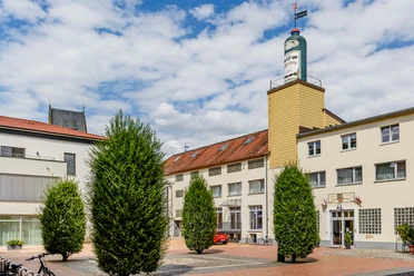 Schlichte Carree (C) pro Wirtschaft GT, Mario Wallenfang Drei hohe, grüne Büsche stehen auf einem gepflasterten Platz vor einem beige-gelben Gebäude mit Turm.