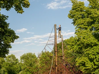 Seilzirkus Eine Person steht auf der Spitze eines hohen roten Klettergerüsts, umgeben von dichtem Grün.