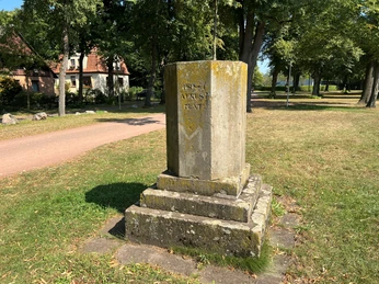 Denkmal aus Stein auf grünem Rasen im Bürgerpark, umgeben von Bäumen und einem rotem Backsteingebäude.