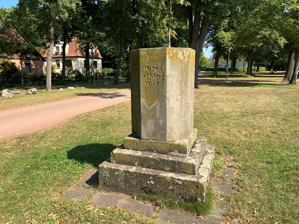 A stone monument on a green lawn in Bürgerpark, surrounded by trees and a red brick building.