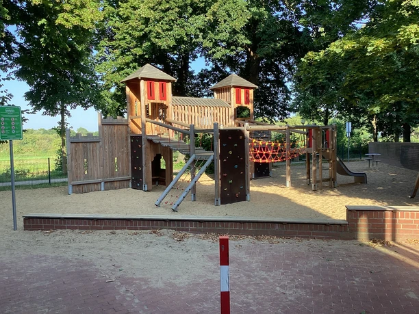 Ein moderner Spielplatz mit Klettergerüst aus Holz, Rutsche und Sandboden, umgeben von Bäumen.