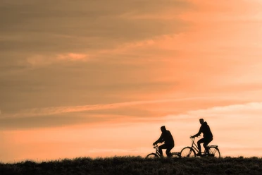 cyclist-3202481_1280.jpg Zwei Radfahrer fahren auf einem Hügel im Abendlicht gegen einen orangefarbenen Himmel.