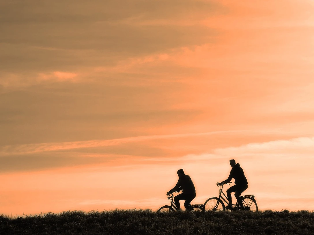 cyclist-3202481_1280.jpg Zwei Radfahrer fahren auf einem Hügel im Abendlicht gegen einen orangefarbenen Himmel.
