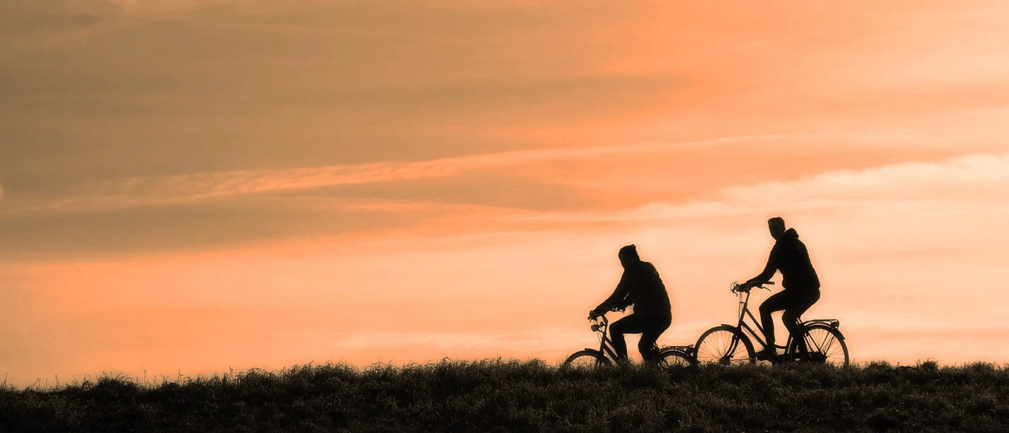 cyclist-3202481_1280.jpg Zwei Radfahrer fahren auf einem Hügel im Abendlicht gegen einen orangefarbenen Himmel.