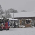 Verschneite Marktstände auf einem Weihnachtsmarkt, umgeben von schneebedeckten Bäumen und Besuchern.