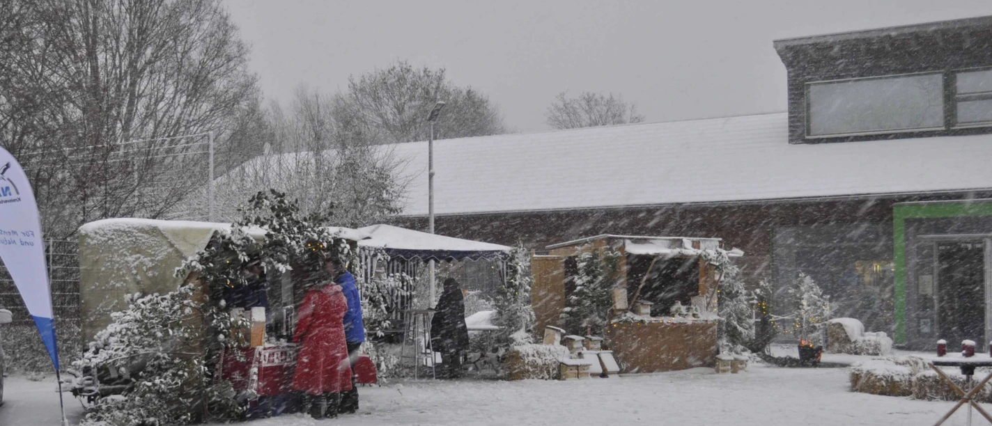 Verschneite Marktstände auf einem Weihnachtsmarkt, umgeben von schneebedeckten Bäumen und Besuchern.