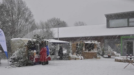 Verschneite Marktstände auf einem Weihnachtsmarkt, umgeben von schneebedeckten Bäumen und Besuchern.