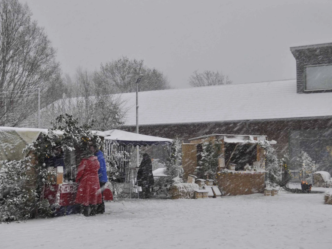 Weihnachtsmarkt Moorhus Verschneite Marktstände auf einem Weihnachtsmarkt, umgeben von schneebedeckten Bäumen und Besuchern.