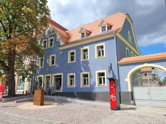 Tourist-Information Bad Lausick mit E-Bike Ladestation - Besucherberatung und Service Außenansicht der Touristinformation Bad Lausick, die Fassade ist leuchtend blau, auf der linken Seite des Eingangs steht eine Kastanie