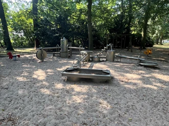 Spielplatz mit Wasserspielen aus Metall, eingerahmt von schattenspendenden Bäumen im Hintergrund.