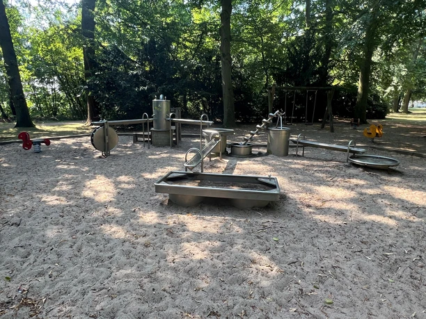 Wasserspielplatz Spielplatz mit Wasserspielen aus Metall, eingerahmt von schattenspendenden Bäumen im Hintergrund.